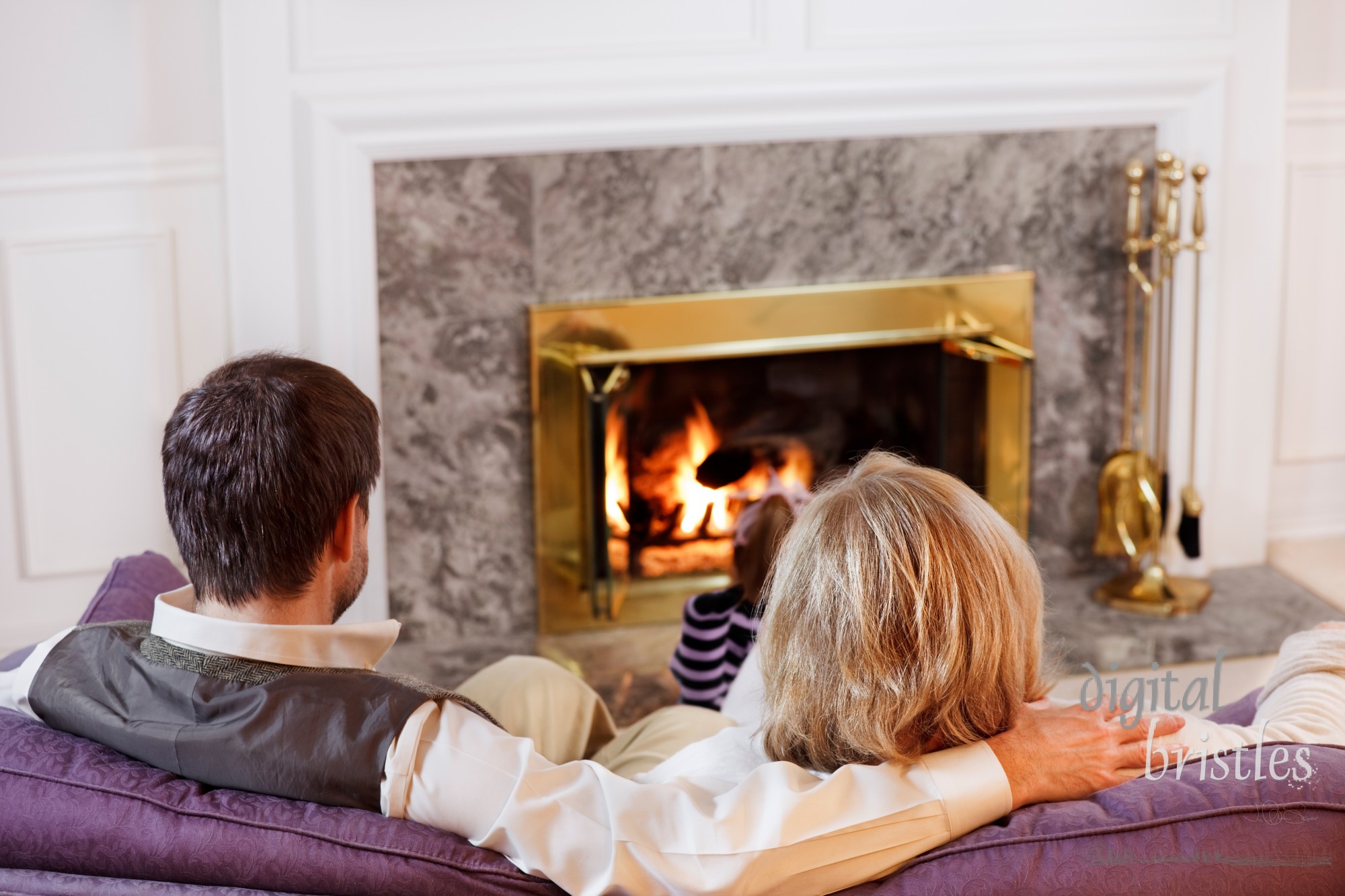 Husband & wife sit together on the sofa while their daughter reads by the fire