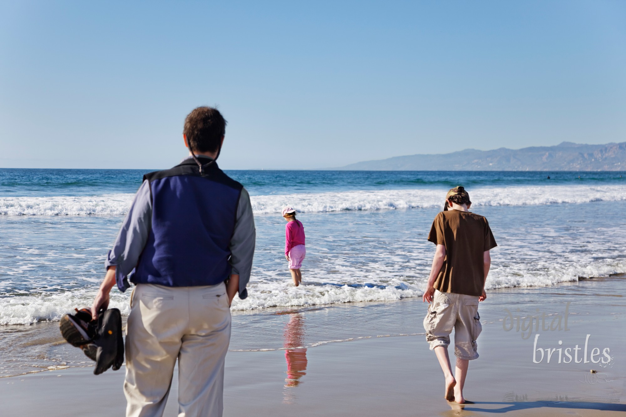 Dad watches his children paddle and play on a sunny winter beach. Focus is on the children & waterline