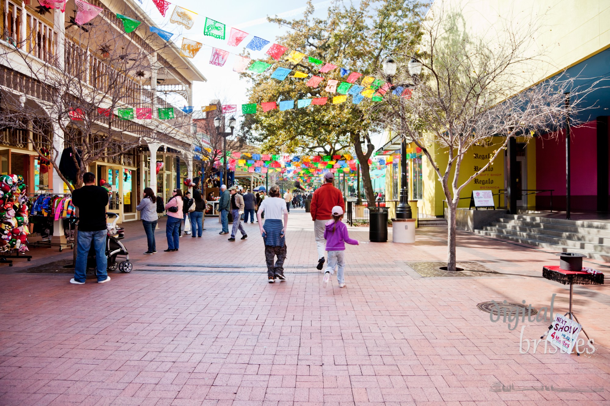 Colorful shops and restaurants line the pedestrian street at the center of Market Square. El Mercado is on the right background and Produce Row shops and La Margarita on the left. Multi-colored papel picado are strung across the walkway. Market Square started as San Antonio's public market in 1894